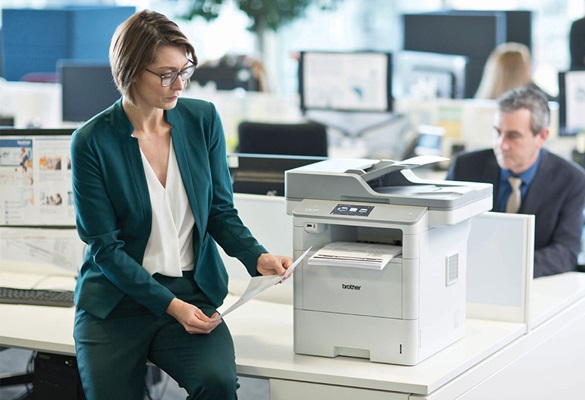 Žena v okuliaroch, v zelenom saku a nohaviciach sediaca na stole vedľa tlačiarne Brother MFC-L6900DW, muž v saku, monitory, stoly